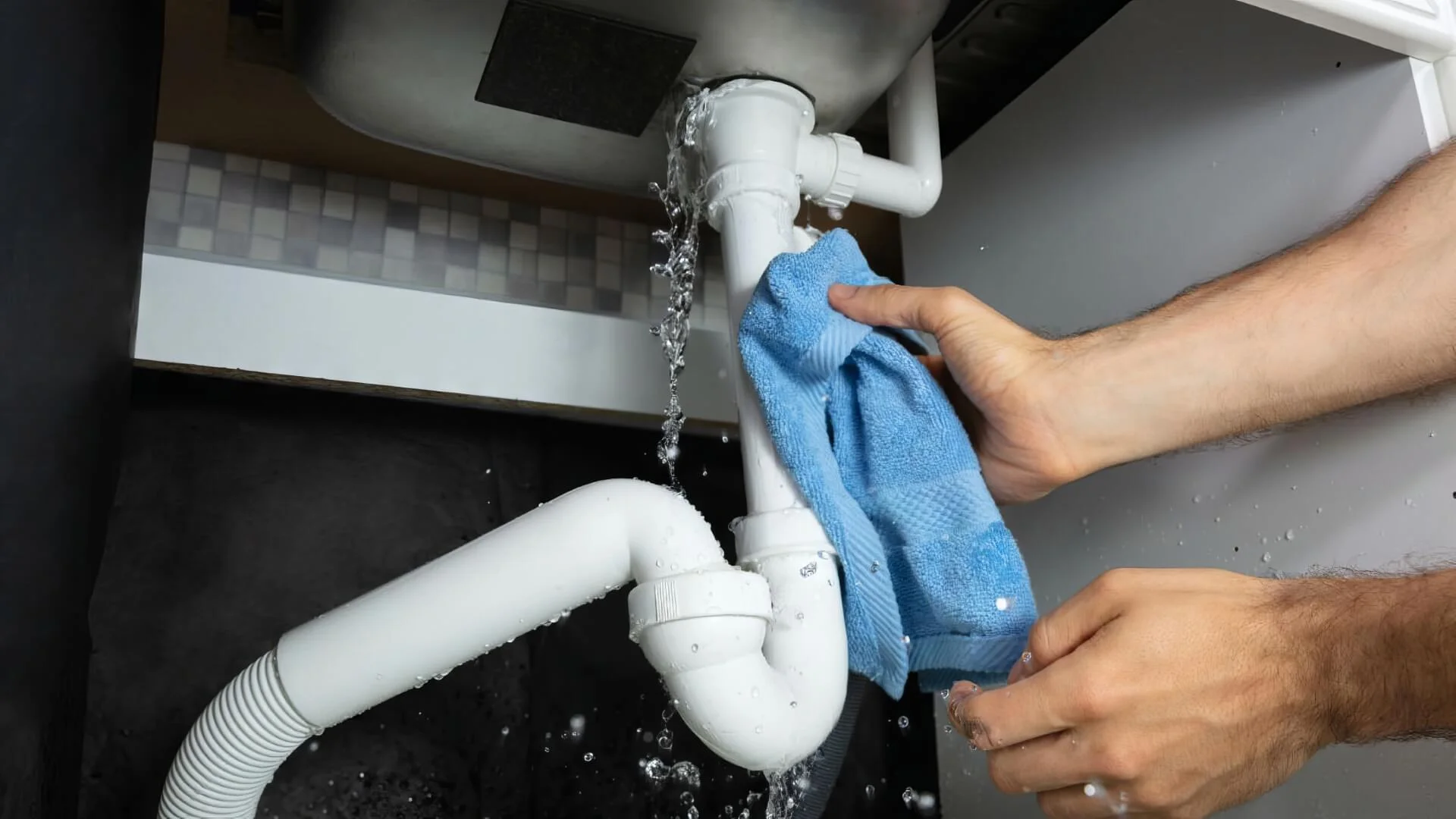 Water leaking from a pipe beneath a sink while a person uses a cloth